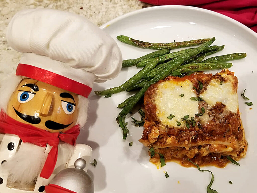 Slice of cheesy lasagna with red meat sauce on a white plate with green beans. There's a nutcracker in the foreground who looks like a chef.