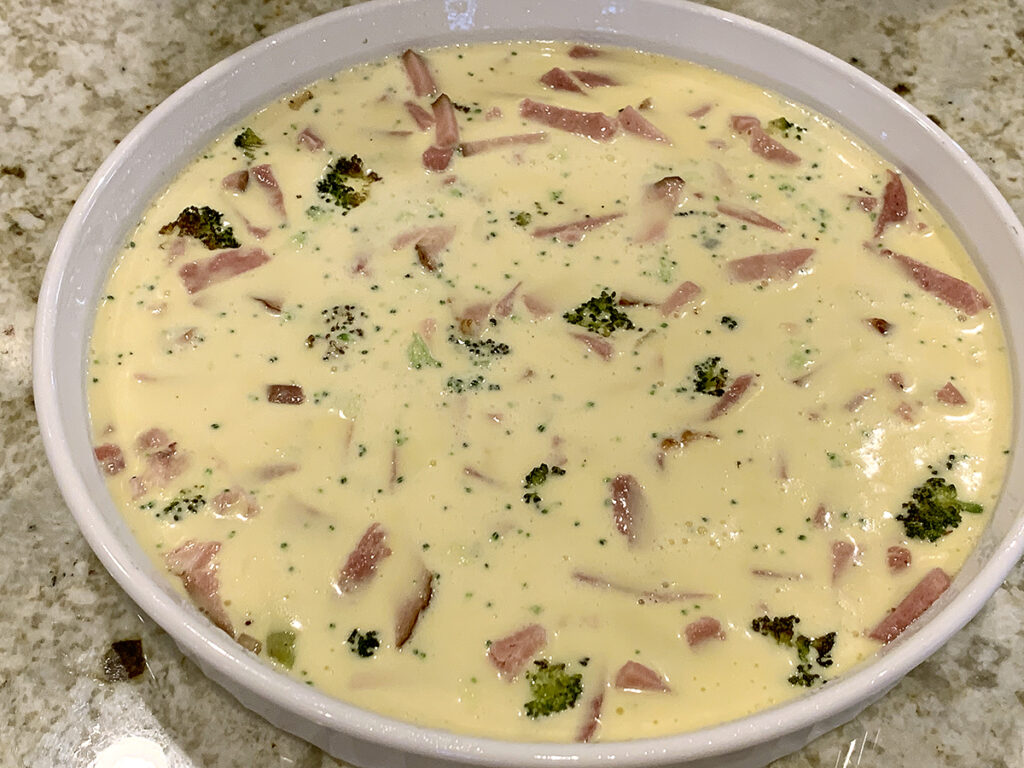 Egg mixture covers ham, broccoli and onions in a white tart pan. 