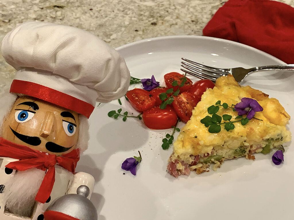 Wedge of ham & broccoli quiche and cherry tomatoes on a white plate garnished with green watercress and purple violets. There's a nutcracker in the foreground who looks like a chef.