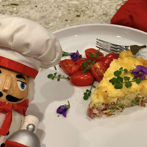 Wedge of ham & broccoli quiche and cherry tomatoes on a white plate garnished with green watercress and purple violets. There's a nutcracker in the foreground who looks like a chef.