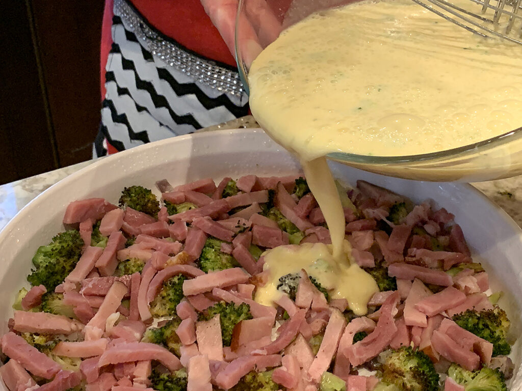 Beaten egg mixture being poured over ham and broccoli in a white tart pan.