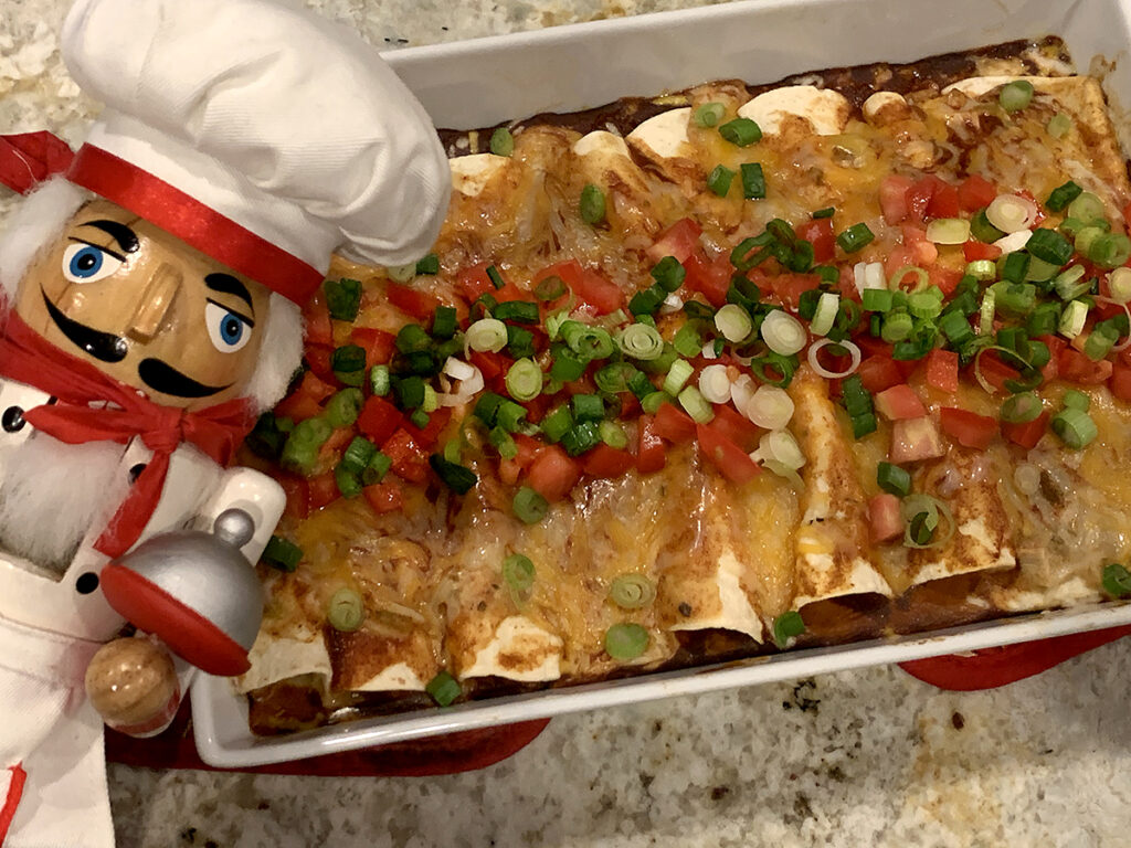 Cheesy and colorful chicken enchiladas in a rectangular white dish. There's a nutcracker in the foreground who looks like a chef.