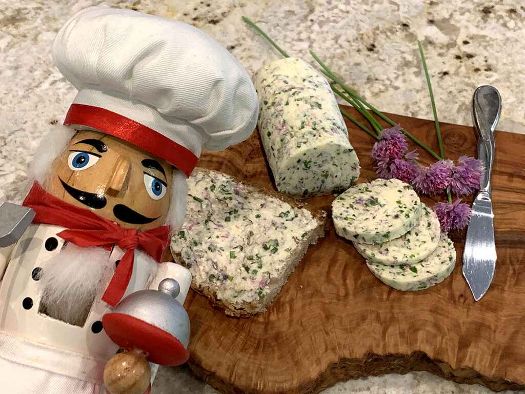 Pale yellow log of butter on a wood board. There are slices of the butter as well as some fresh chives with pink blossoms and more butter spread on a piece of bread. There's a nutcracker in the foreground who looks like a chef.