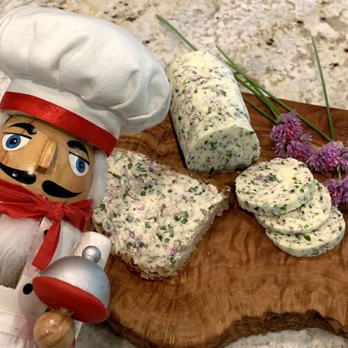 Pale yellow log of butter on a wood board. There are slices of the butter as well as some fresh chives with pink blossoms and more butter spread on a piece of bread. There's a nutcracker in the foreground who looks like a chef.