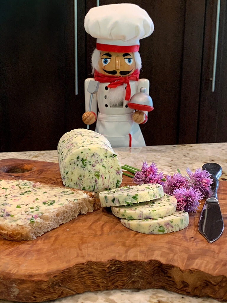 Pale yellow log of butter on a wood board. There are slices of the butter as well as some fresh chives with pink blossoms and more butter spread on a piece of bread. There's a nutcracker in the background who looks like a chef.