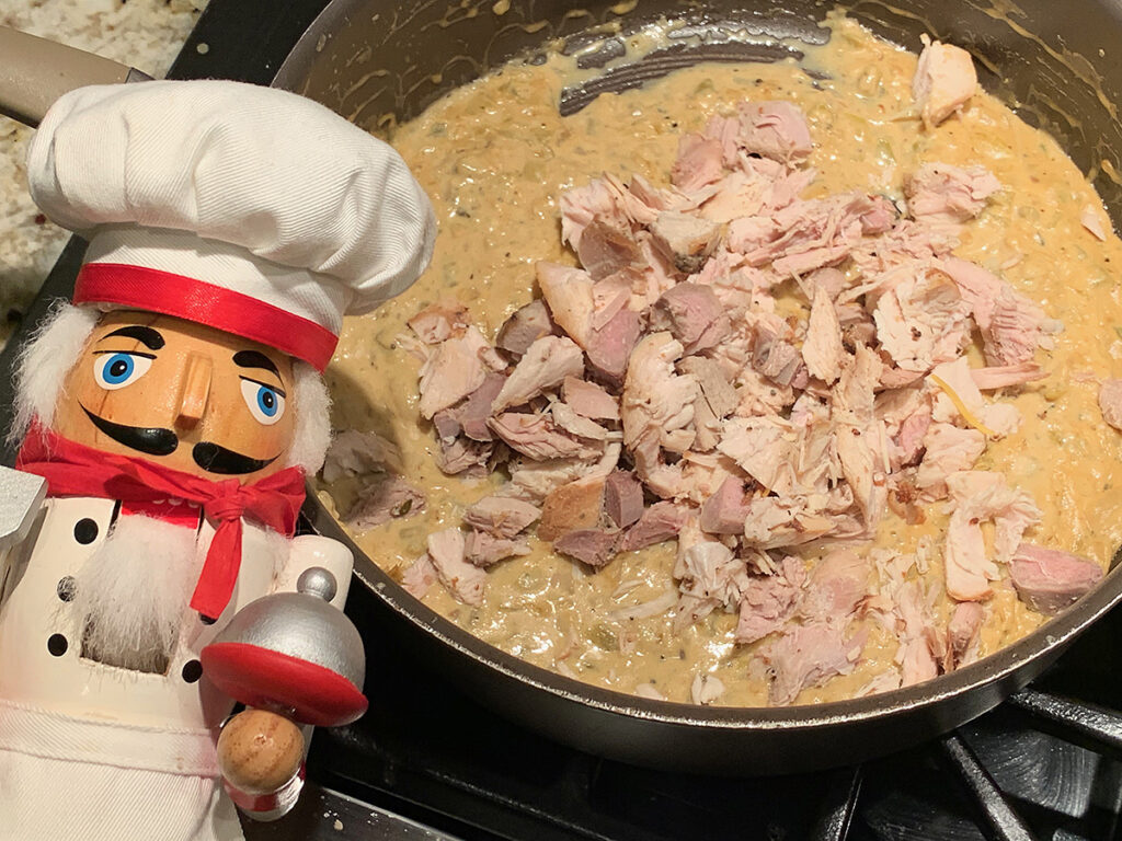 Cooked cut up chicken added to a skillet of white sauce. There's a nutcracker in the foreground who looks like a chef.