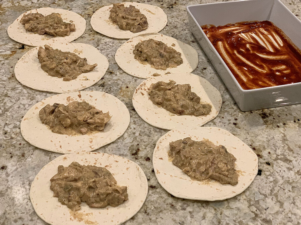 Chicken enchilada filling divided amongst 8 flour tortillas, red sauce spread on the bottom of a white casserole dish. 