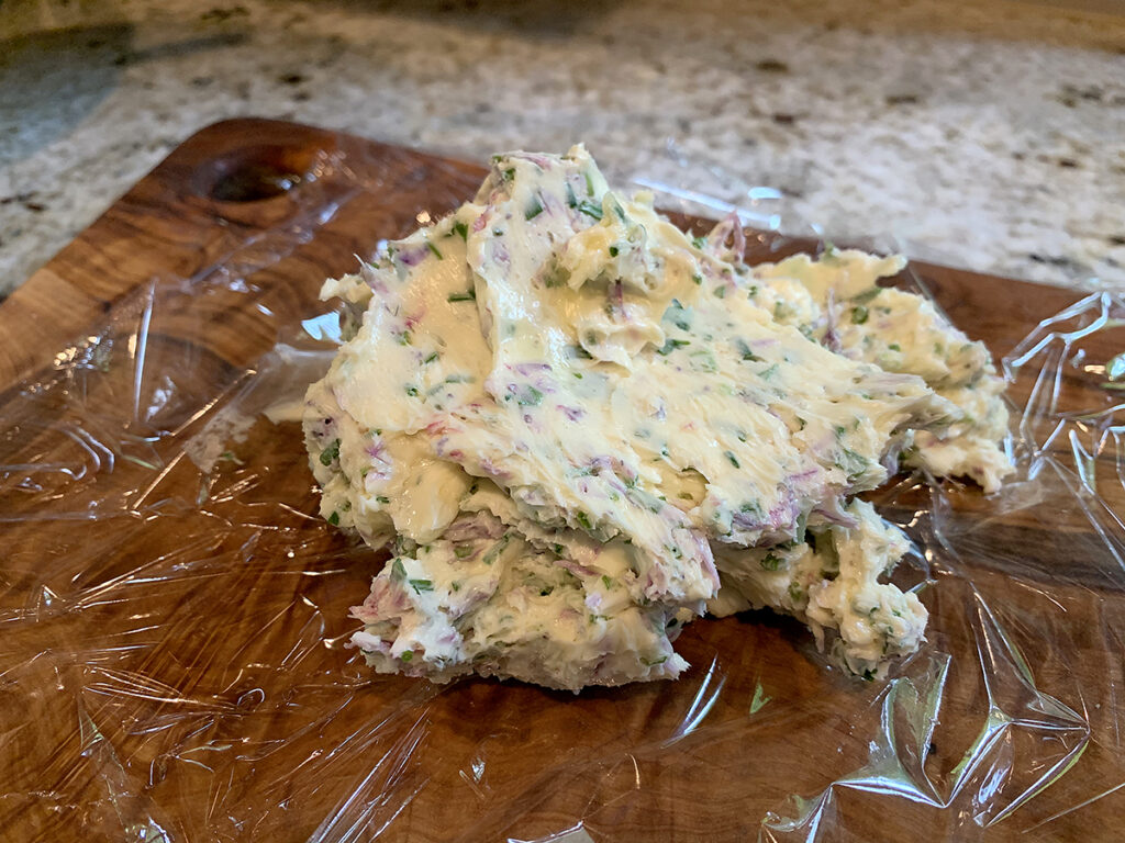 A mound of butter with pink and green flecks laying on a piece of cling film, on a board.
