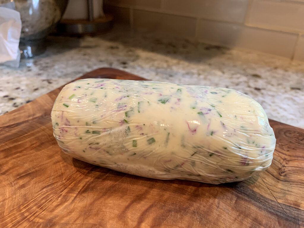 Pale yellow log of butter with pink and green flecks on a wood board.