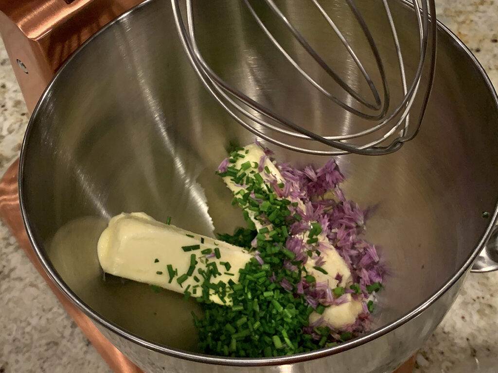 Two sticks of butter, minced green chives and pink chive blossoms in a steel bowl of the stand mixer.