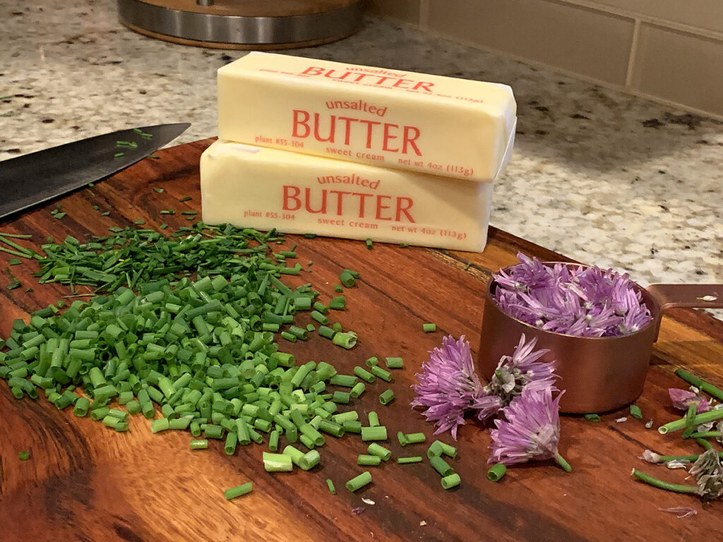Two sticks of butter, minced green chives and pink chive blossoms on a wood board.