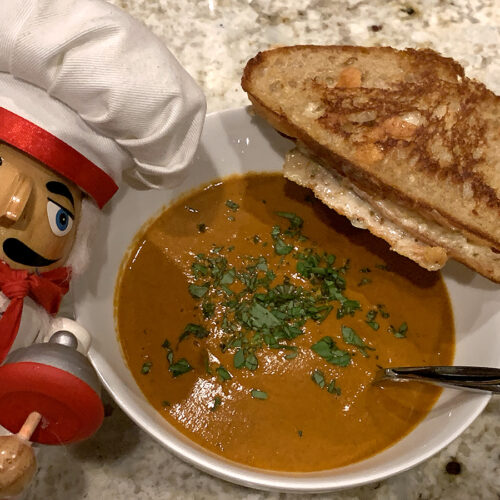 Rustic grilled cheese with a bowl of homemade tomato soup on a wood board. There's a nutcracker that looks like a chef in the foreground.