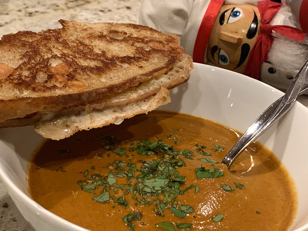 Rustic grilled cheese with a bowl of homemade tomato soup on a wood board. There's a nutcracker that looks like a chef in the foreground.