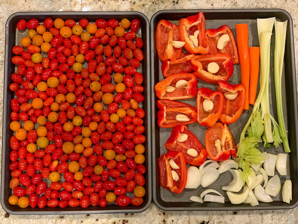 Two sheet pans – one filled with red and yellow grape tomatoes, the other with bell peppers, garlic, onions, carrots and celery.
