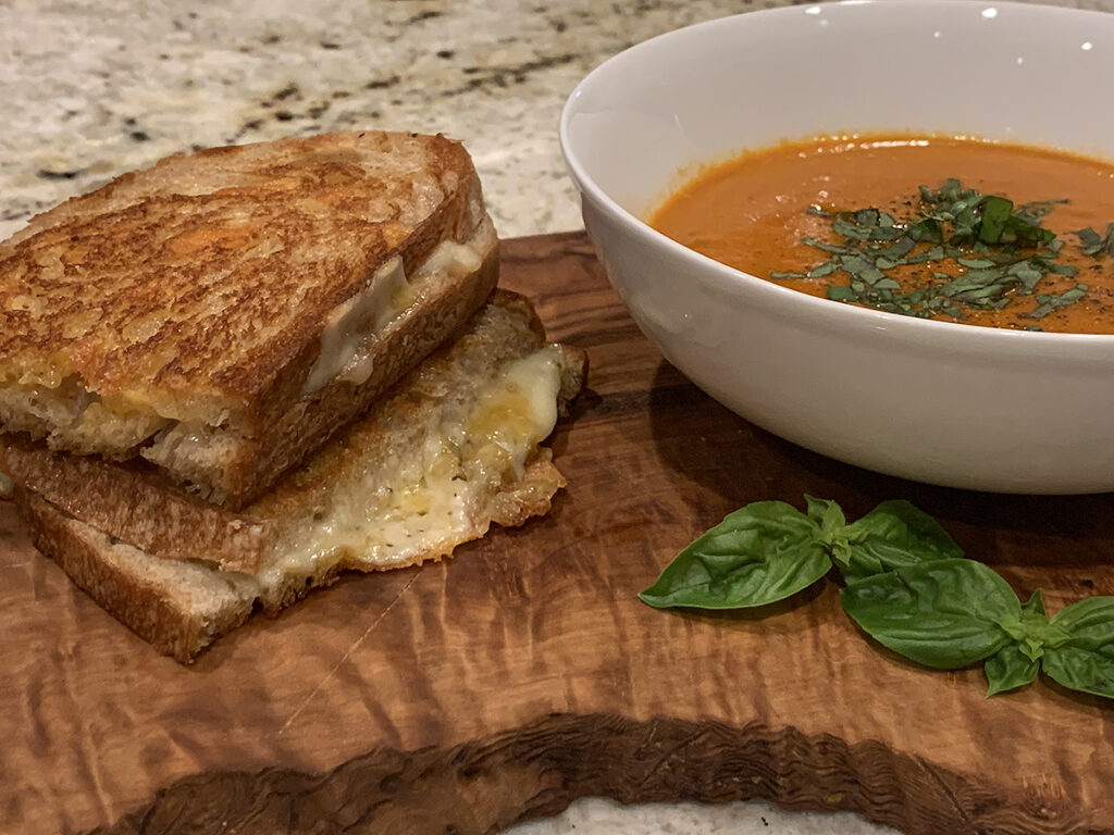 Rustic grilled cheese with a bowl of homemade tomato soup on a wood board.