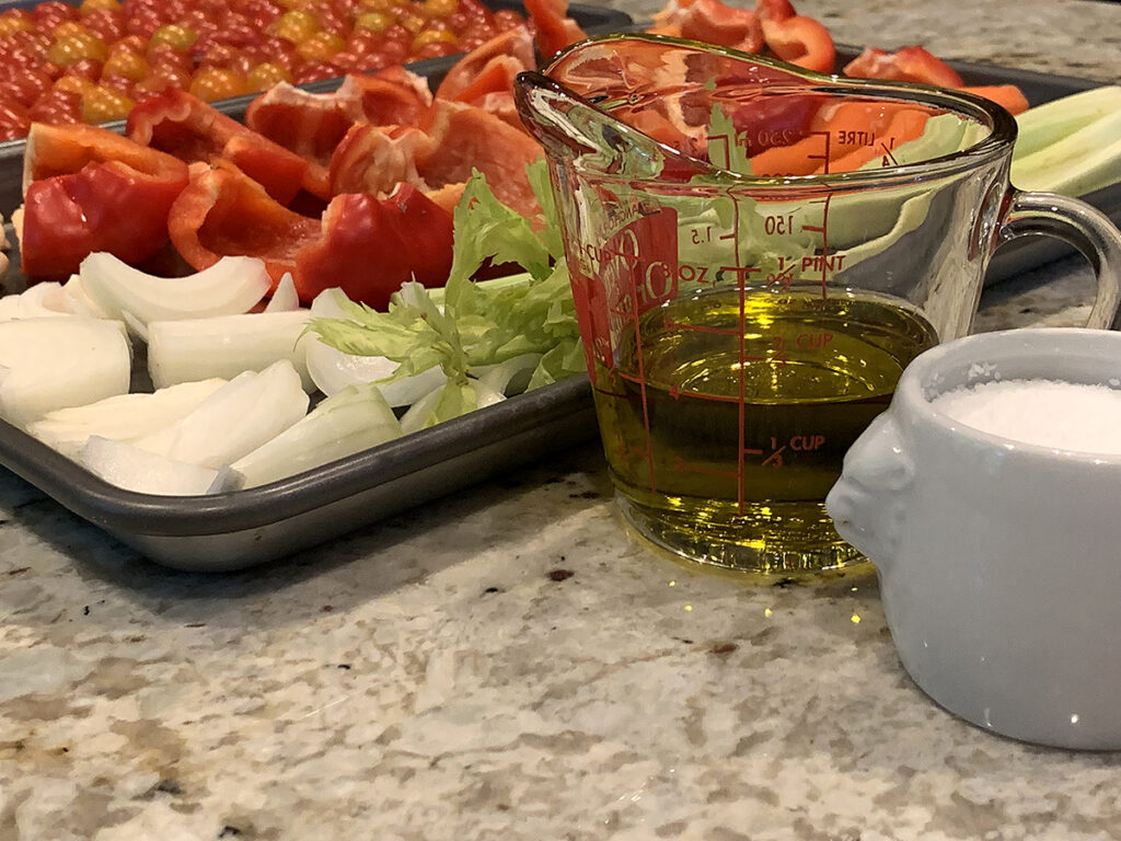 close up of veggies on a sheet pan with olive oil in a measuring cup and kosher salt in a white ceramic dish.