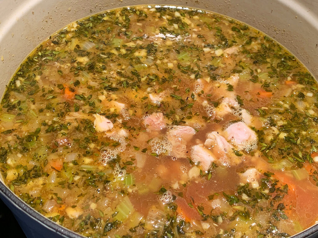 Brown soup broth with lots of herbs, carrots, chunks of poultry in a white dutch oven.