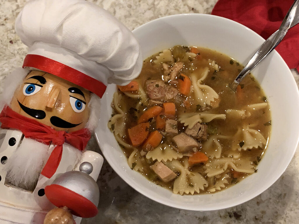 Brown soup broth with butterfly pasta, carrots, chunks of poultry in a white bowl. There's a nutcracker who looks like a chef in the foreground.