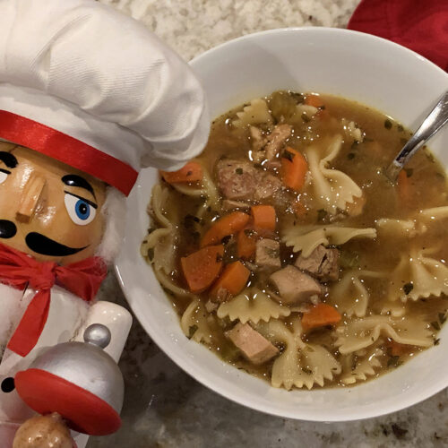Brown soup broth with butterfly pasta, carrots, chunks of poultry in a white bowl. There's a nutcracker who looks like a chef in the foreground.