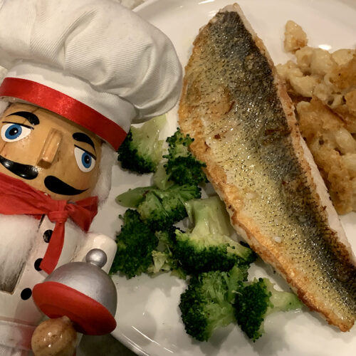 Pan Seared Walleye, skin side up on a white plate with broccoli and macaroni and cheese. There's a nutcracker in the foreground who looks like a chef.
