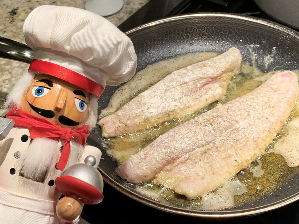 Two long pink fish fillets that were dredge in flour laying in a grey skillet. There's a nutcracker in the foreground who looks like a chef.