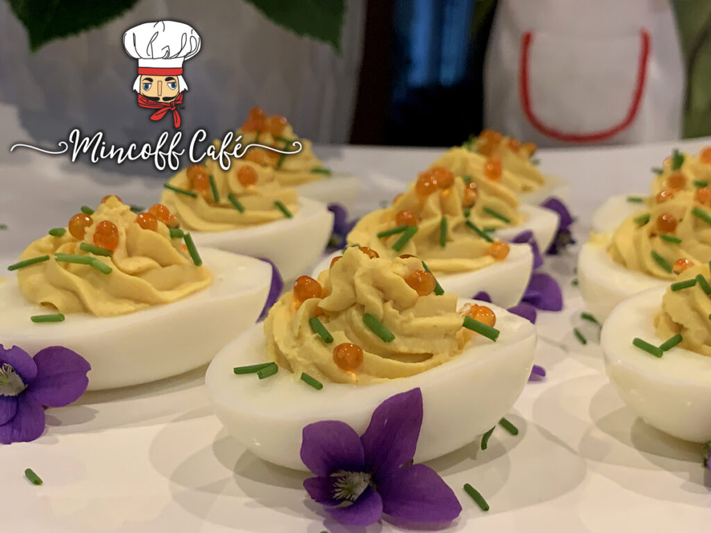 Close up of beautiful deviled eggs on a white platter garnished with orange caviar, green chives and purple violets.