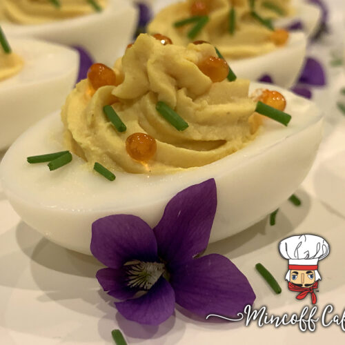 Close up of beautiful deviled eggs on a white platter garnished with orange caviar, green chives and purple violets.