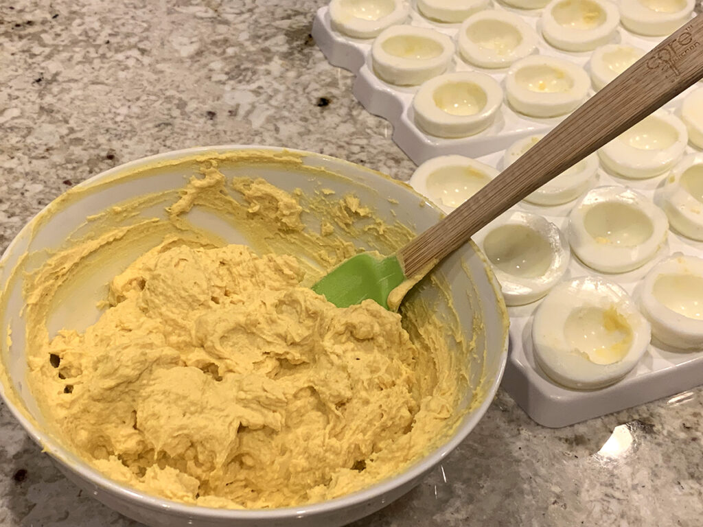 Deviled egg mixture in a white bowl ready to start piping into the hard boiled egg halves to the right.
