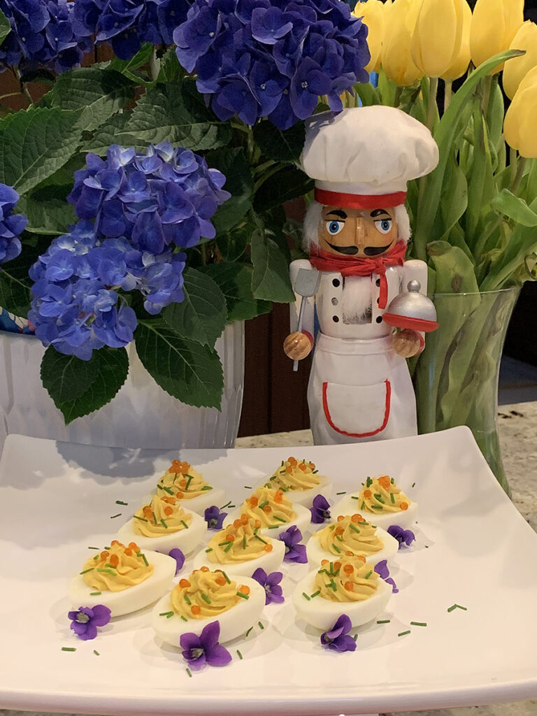 Beautiful deviled eggs on a white platter garnished with orange caviar, green chives and purple violets. There'a nutcracker in the background who looks like a chef.