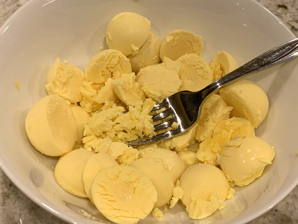 several cooked egg yolks in a white bowl, being mashed with a fork.