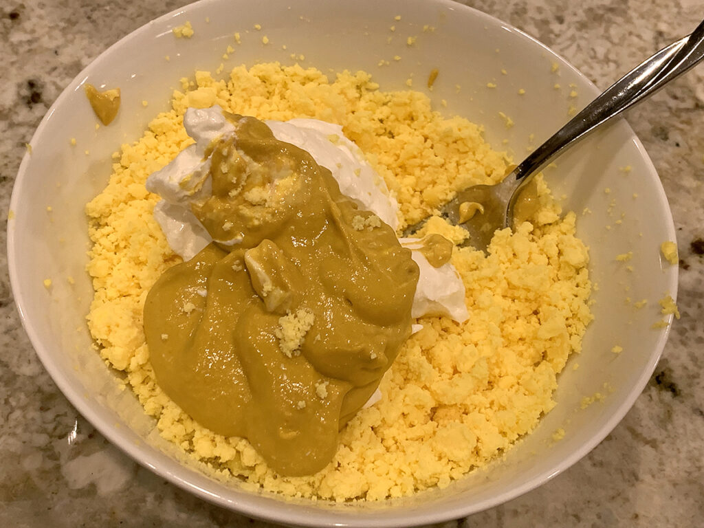 Mashed up hard boiled egg yolks in a white bowl with yellow mustard and yogurt. Ready to mix.