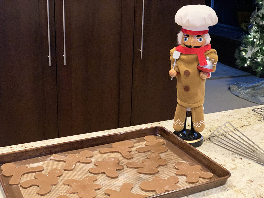 A sheet pan full of gingerbread men cookies ready to go in the oven. There's a nutcracker who looks like a chef, wearing a gingerbread man costume looking over them.