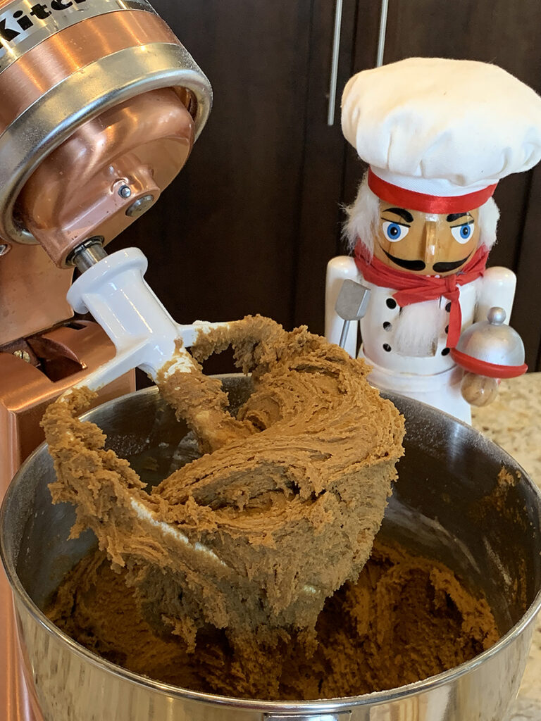 Brown gingerbread dough with a somewhat wet consistency in a stand mixer with a paddle attachement. There's a nutcracker in the background who looks like a chef.