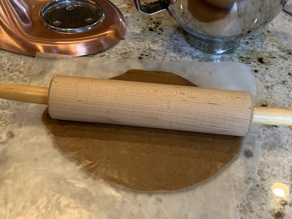 Gingerbread cookie dough sandwiched between pieces of wax paper rolled out with a rolling pin on top.