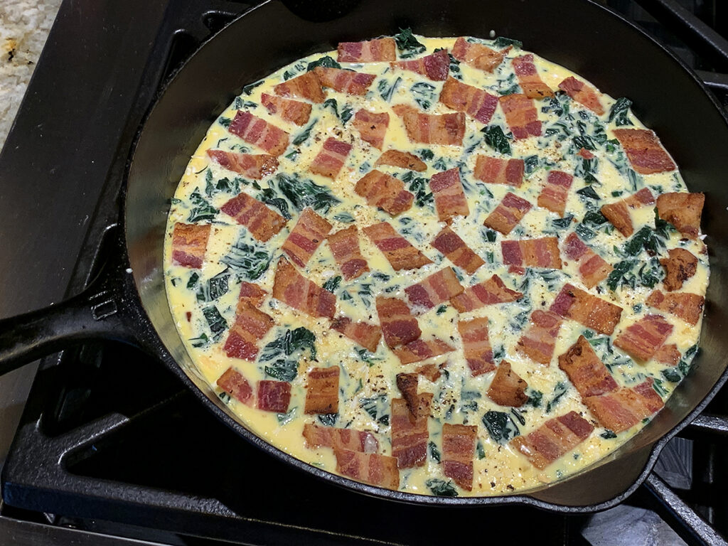 Beaten eggs in an iron skillet with chopped green stinging nettles and chopped bacon layered on top.