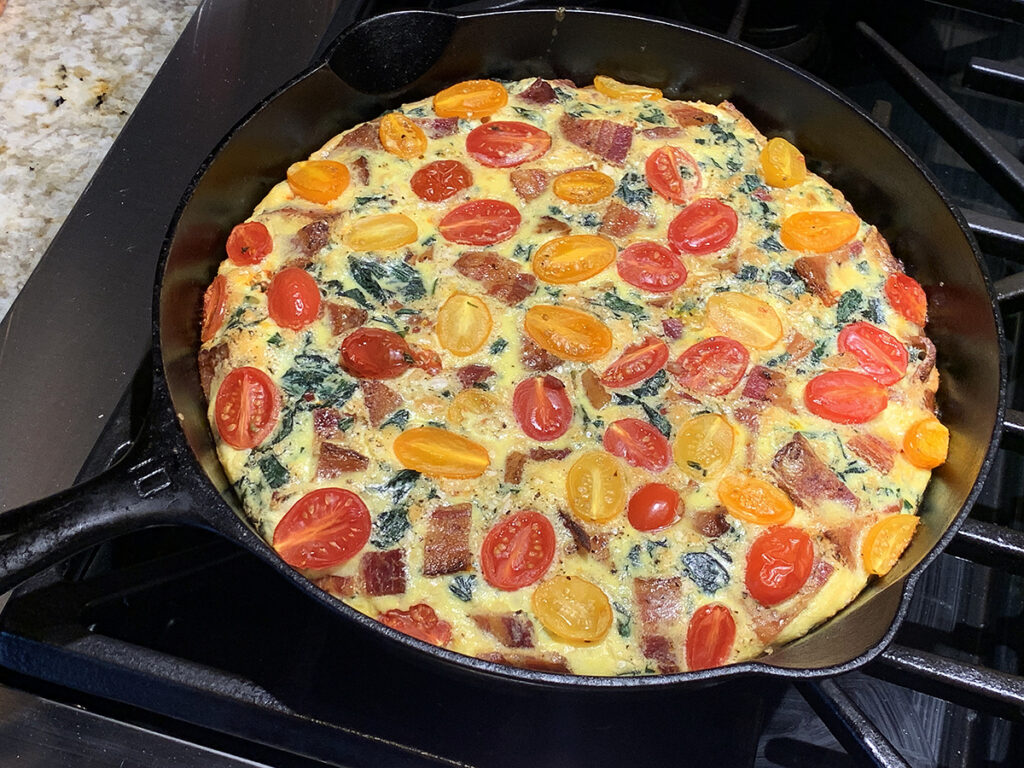 Cooked beaten eggs in an iron skillet with chopped green stinging nettles, chopped bacon and colorful cherry tomatoes layered on top.