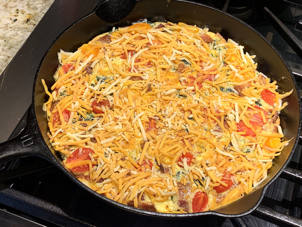 Baked egg frittata in an iron skillet layered with shredded cheddar cheese ready for the oven.