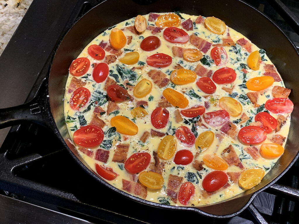 Beaten eggs in an iron skillet with chopped green stinging nettles, chopped bacon and colorful cherry tomatoes layered on top.