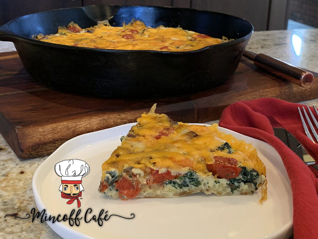 A wedge of a bacon tomato frittata with a hash brown crust on a white plate. The rest is in an iron skillet in the background.