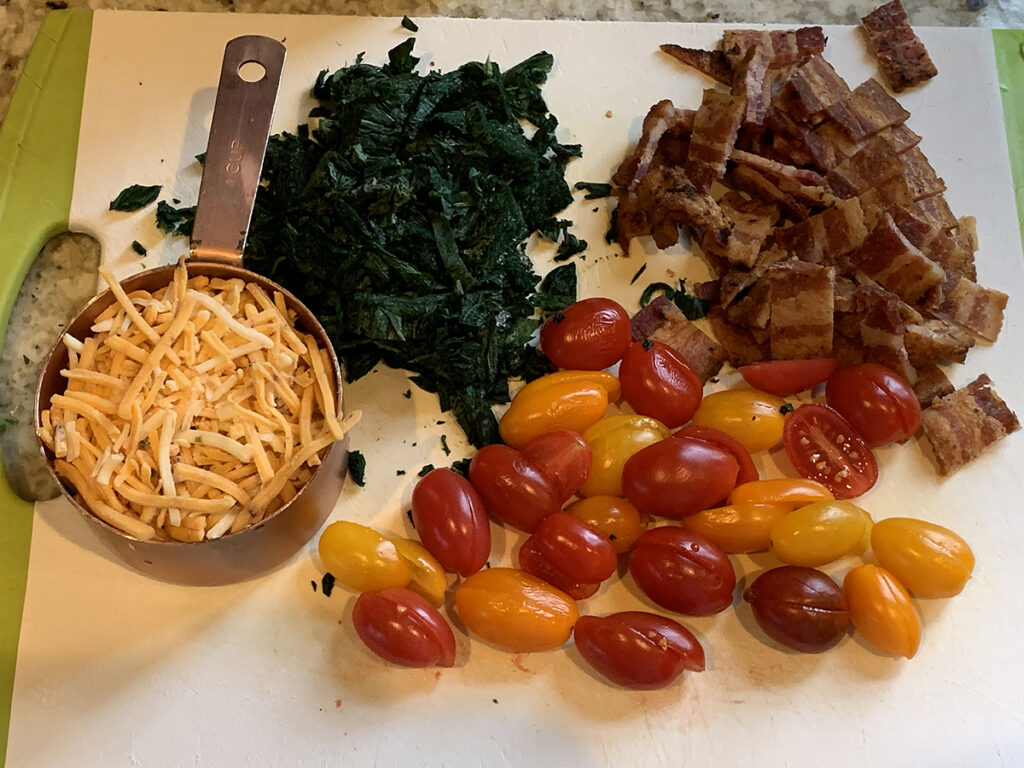 Shredded cheddar cheese in a copper measuring cup, colorful cherry tomatoes cut in halves, chopped cooked bacon, chopped cooked stinging nettles on a white cutting board.